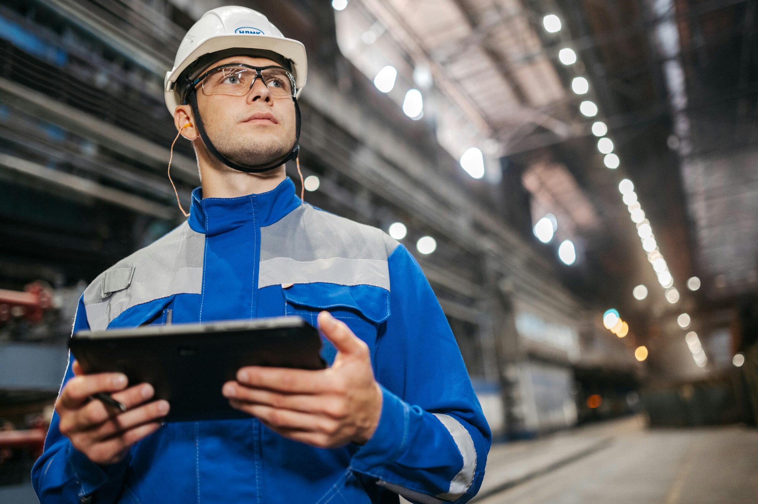 pexels-photo-32845692-32845692 Factory worker in safety gear using a tablet in a well-lit industrial setting.