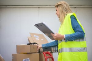 Woman in high-visibility vest handling boxes for delivery service inside a warehouse setting.