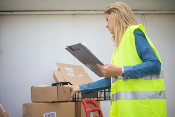 Woman in high-visibility vest handling boxes for delivery service inside a warehouse setting.