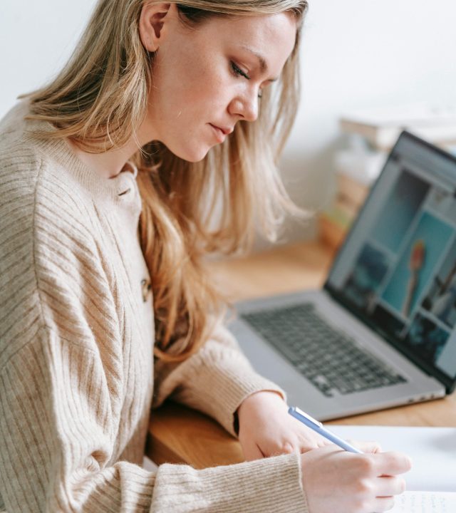 Side view of crop focused female taking notes in planner at table with netbook on blurred background
